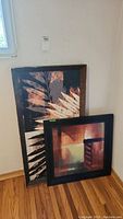 Two framed prints leaning against wall, one larger vertical leaf design with chipped frame, other smaller square color block print with scratched frame.