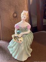 Front view of Royal Doulton Clarissa porcelain figurine showing seated young woman in green dress with pink sleeves holding basket of flowers