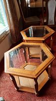 Two hexagonal wooden side tables with beveled glass tops and lower shelves shown from above and angled side view on carpeted floor near window and dining chairs.