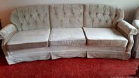 Front view of beige three-seat tufted sofa with arm covers and skirted base showing fabric upholstery and cushion details.