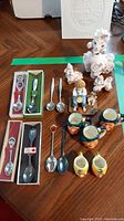 Photo showing all eight souvenir teaspoons, three poodle figurines, ceramic boy with dog figurine, and five miniature 'head' mugs all arranged on a wooden surface.