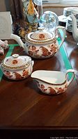 Three-piece Wedgwood Golden Cockerel tea set on wooden table, includes teapot, sugar bowl with lid, and creamer, decorated with gold and rust floral and rooster patterns.