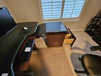 View showing L-shaped black computer desk with a wood finished 2 drawer collapsible credenza underneath window, keyboard and mouse on desk.
