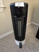 Front view of De'Longhi ceramic tower heater standing on carpet with remote placed in front of it. Visible control buttons and LED indicator light on top of the unit.