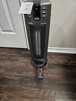 Black De'Longhi ceramic tower heater standing upright on wood floor next to a white door, with remote control placed in front of heater base.