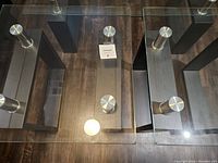 Top view of two matching glass end tables with black wood bases and silver metal supports on a wood floor.