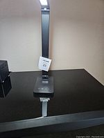 Black metal LED desk lamp standing on black surface with visible touch control panel and adjustable arm, lamp is lit in white LED color.