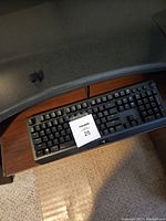 Black Razer BlackWidow Elite mechanical keyboard on a wooden desk surface. Keyboard has standard QWERTY layout, media controls and volume dial on top right, and magnetic wrist rest attached at bottom.