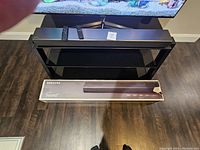 Front view of Samsung soundbar placed on a black glass stand with two remotes and original box.