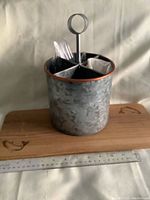 Metal cutlery caddy with four sections, central handle ring, holding some plastic utensils and napkins, placed on a cedar plank with engraved fish decorations and a ruler for size reference.