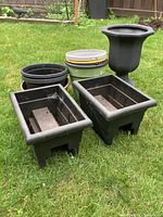 Black rectangular balcony planters, round pots stacked and an ornate urn on grass