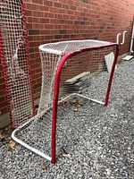 Photo of a 6ft x 4ft hockey net with red metal frame and white netting showing wear, dents, and holes in the net. Two stray puck panels leaning against the wall beside the net.