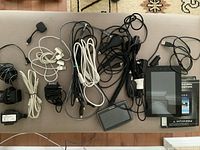 Top-down view of various cords and electronic items including USB cables, adapters, a black GPS or similar device, and the Nextbook Premium 7 tablet with manual.