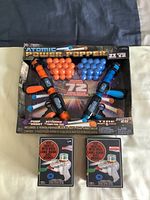 Photo showing the Atomic Power Popper set including two pump-action launchers with orange and blue parts and 72 soft foam balls in matching colors. Also visible are two boxed Disc Shooters with a multi-colored disc holder.