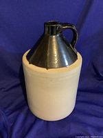 Front angle view of gallon stoneware whisky jug showing black glazed top with handle, cream colored base, and small chip on rim.