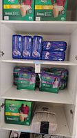 Wide view of a shelf displaying boxes and packs of adult protection underwear: green boxes of Depend Protection Plus Ultimate, blue packs of Tena Ultimate Plus, and purple packs of Depend Night Defense, with bundles of folded white Depend underwear.