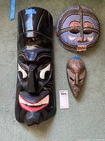 Three African wood carved masks arranged on carpet: one large black mask, one round colorful mask, and one smaller elongated bronze-like mask.