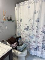 Full view of butterfly-themed shower curtain, stacked towels in various colors on toilet, stainless steel trash can visible on top towel, and reed diffuser on glass shelf