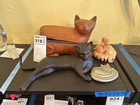 Multiple cat figurines arranged together on a shelf including wooden, metal, and carved wood cats.