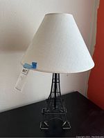 Photo showing the lamp with its metal Eiffel Tower shaped base and white fabric shade on top, with a tag showing lot number and price.