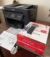 Black Epson WorkForce Pro WF-3820 printer on wooden table with instruction papers and two sealed reams of copy paper and an Epson 822 black ink cartridge.