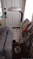 Full view of Bell and Howell white plastic flexible floor lamp standing beside couch and magazine rack in front of door, showing round base, flexible neck, and dome-shaped head.