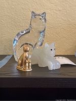 Three cat figurines placed on a dark surface; clear crystal, white marble, and gold-tone metal Egyptian cat with smaller kitten