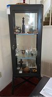 Front view of the Ikea Fabrikor 3-tier metal curio cabinet showing glass doors, interior shelves, included key in lock, and various items displayed inside (not included)