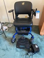 Front view of the Golden LiteRider Envy power chair showing black cushioned seat, armrests, joystick control, footrest, and charger on carpeted floor.