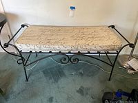 Metal bench with black curved and scroll metal frame, beige cushioned seat with cursive script print. Shown on blue carpet with electrical cords nearby.