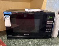 Front view of black Panasonic microwave model NN-SN651B with digital keypad and display on a kitchen countertop with a paper towel roll nearby.