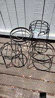 Four black metal round plant pot stands arranged near a white wall on wooden decking, showing overall condition and design