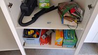 Photo of upper and lower shelves in white cabinet showing black shop vacuum, green shop bag, Swiffer pads, extension cords, gloves, hammer, and plastic tool container.