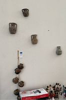 Five wall-mounted small pottery vessels and a stringed wall hanging with six small pottery pots arranged vertically on a white wall.