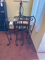 Pair of wrought iron plant tables with round glass tops, placed indoors on carpet near window. One glass top has visible scratches.