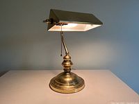 Front view of the vintage brass bankers desk lamp on a white surface against gray background, light turned on, showing pull chain and detail of base and shade.