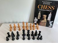 Two complete sets of chess pieces arranged on a table, one light wood and one dark wood, with the chess book standing behind them.