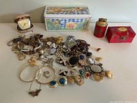 Photo showing the complete lot of various 1980s costume jewelry including earrings, necklaces, bracelets, brooches, and rings displayed on a white surface with decorative tins and a clock in the background