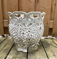 Front angled view showcasing the intricate lattice and floral cut-out pattern of the white painted metal planter with some rust spots and weathering on a wooden surface.