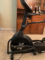 Side view showing elliptical trainer with display console, pedals, and handles, on carpet in front of wooden cabinet.