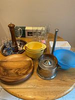 Photo showing a variety of kitchen items arranged on a wooden table, including yellow mixing bowls, wooden pepper grinder, large wooden bowl, stainless steel teapot, blue colander, beer glasses, and white ceramic napkin holder.