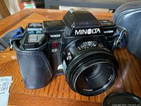 Front-side angle showing black Minolta Maxxum 7000 camera body with attached 50mm AF lens, lens cap off, placed on wooden table with blue camera strap attached.