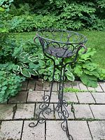 Full view of black wrought iron planter with decorative circular pattern and tripod curved leg stand set outdoors on stone pavers with garden foliage background.