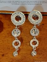 Front view of the pair of silver-tone drop earrings with clear Swarovski crystals arranged in linked circular segments: large hollow crystal-encrusted circle, bezel-set round crystal, smaller crystal circle, and bottom crystal drop.