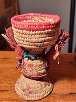 Front view of woven basket woman figure with red rim basket, turquoise necklace, and woven feathers on arms.