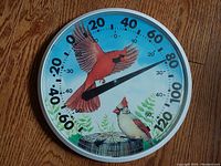 Front-facing image of circular thermometer featuring two cardinals, Fahrenheit scale, and blue sky background with foliage and post illustration.
