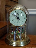 Full front view of glass dome clock showing clock face and miniature dancers inside, gold-tone base and decorative scrollwork above clock face.
