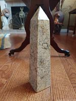 Front view of the vintage stone Egyptian obelisk sculpture on wooden floor with furniture in background, showing its pyramid-shaped top and tapered four-sided design.