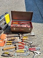 Vintage metal Sears Craftsman toolbox with an assortment of used hand tools including hammers, pliers, wrenches, wire cutters, measuring tape, padded knee pads spread on gravel surface.