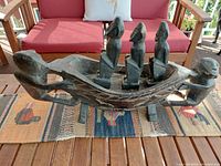 Front and side view of the entire wooden canoe carving with three figures inside and two on exterior, placed on a wooden table with red cushioned chairs in the background.